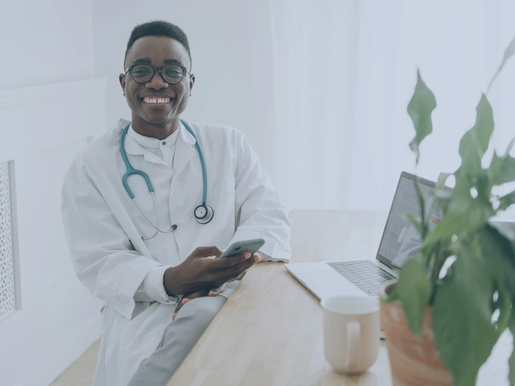Digitale Reifegradmessung Krankenhäuser Ein Arzt sitzt am Schreibtisch, in der Hand ein Mobiltelefon, vor sich einen Laptop. Er schaut uns an.
