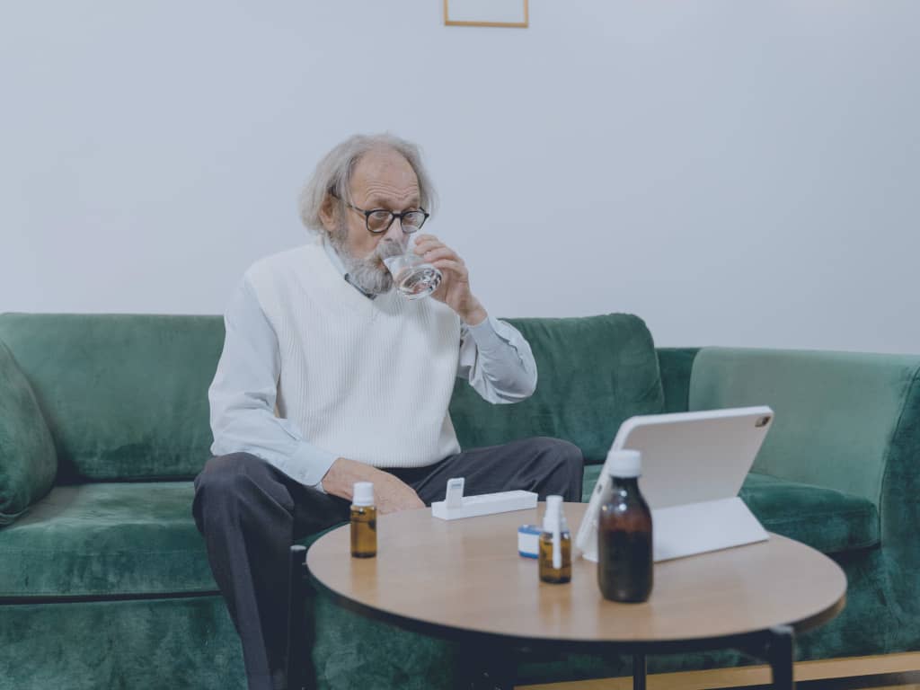 Ein älterer Patient sitzt auf seiner Couch, nimmt gerade Medikamente ein, während er auf seinem Pad einen Arzt konsultiert.
