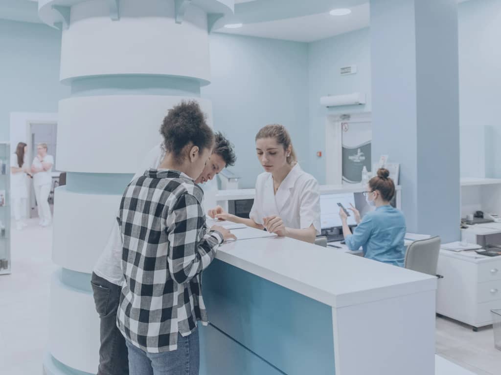 Eine Patientin und ein Patient füllen in der klinischen Anmeldung Formulare aus. Eine Mitarbeiterin steht hinter dem Tresen und hilft.