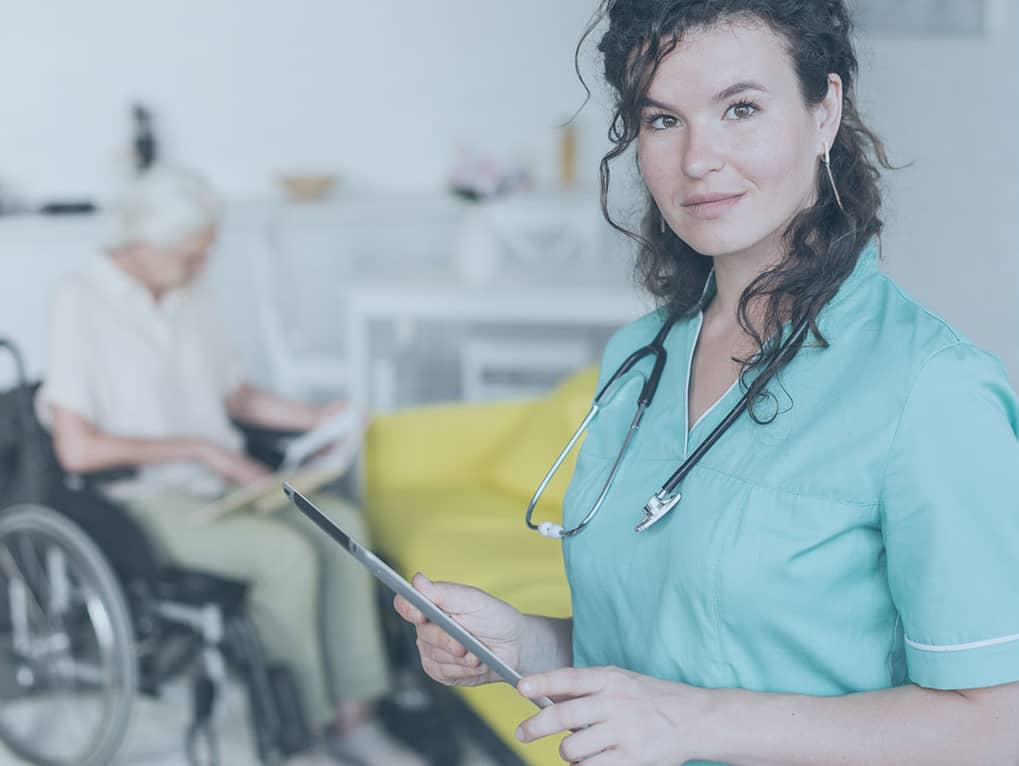 Eine Pflegekraft hält ein Pad. Im Hintergrund sitzt eine Dame im Rollstuhl.