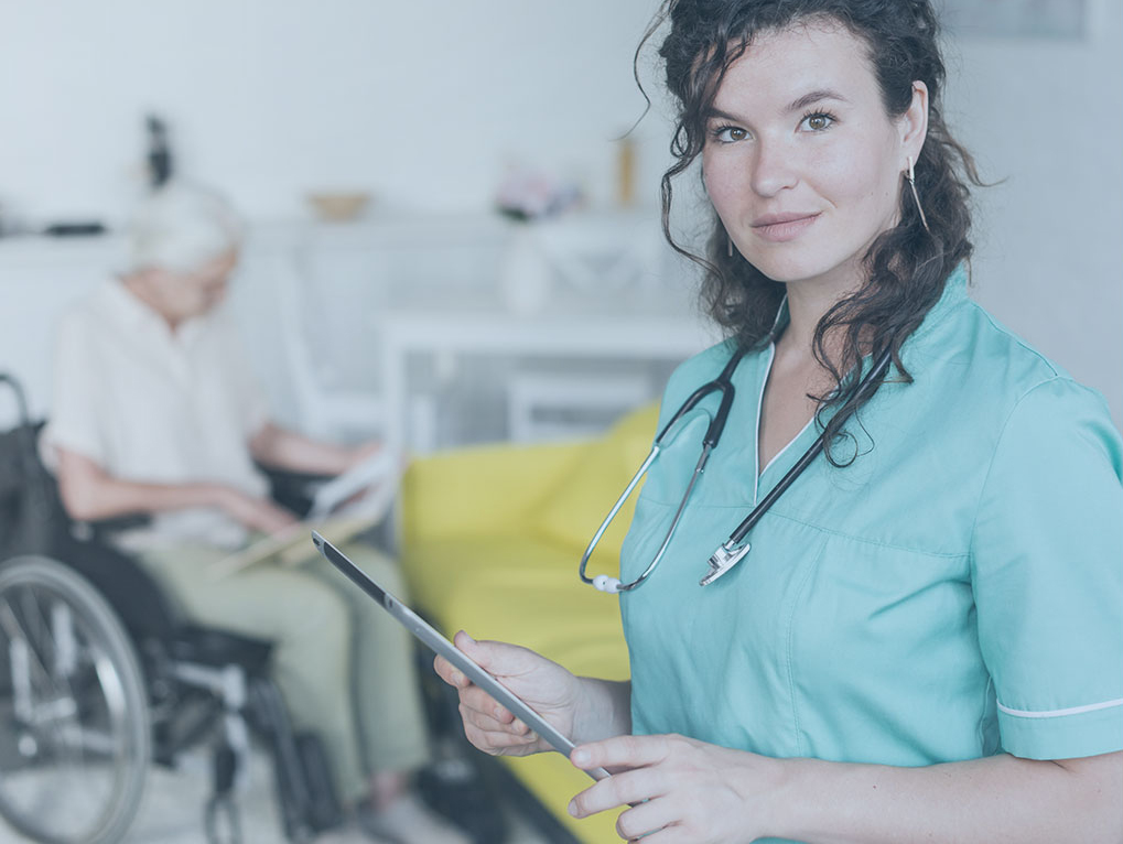 ehealth gesetz und digitale Alternativen Eine Pflegekraft hält ein Pad. Im Hintergrund sitzt eine Dame im Rollstuhl.