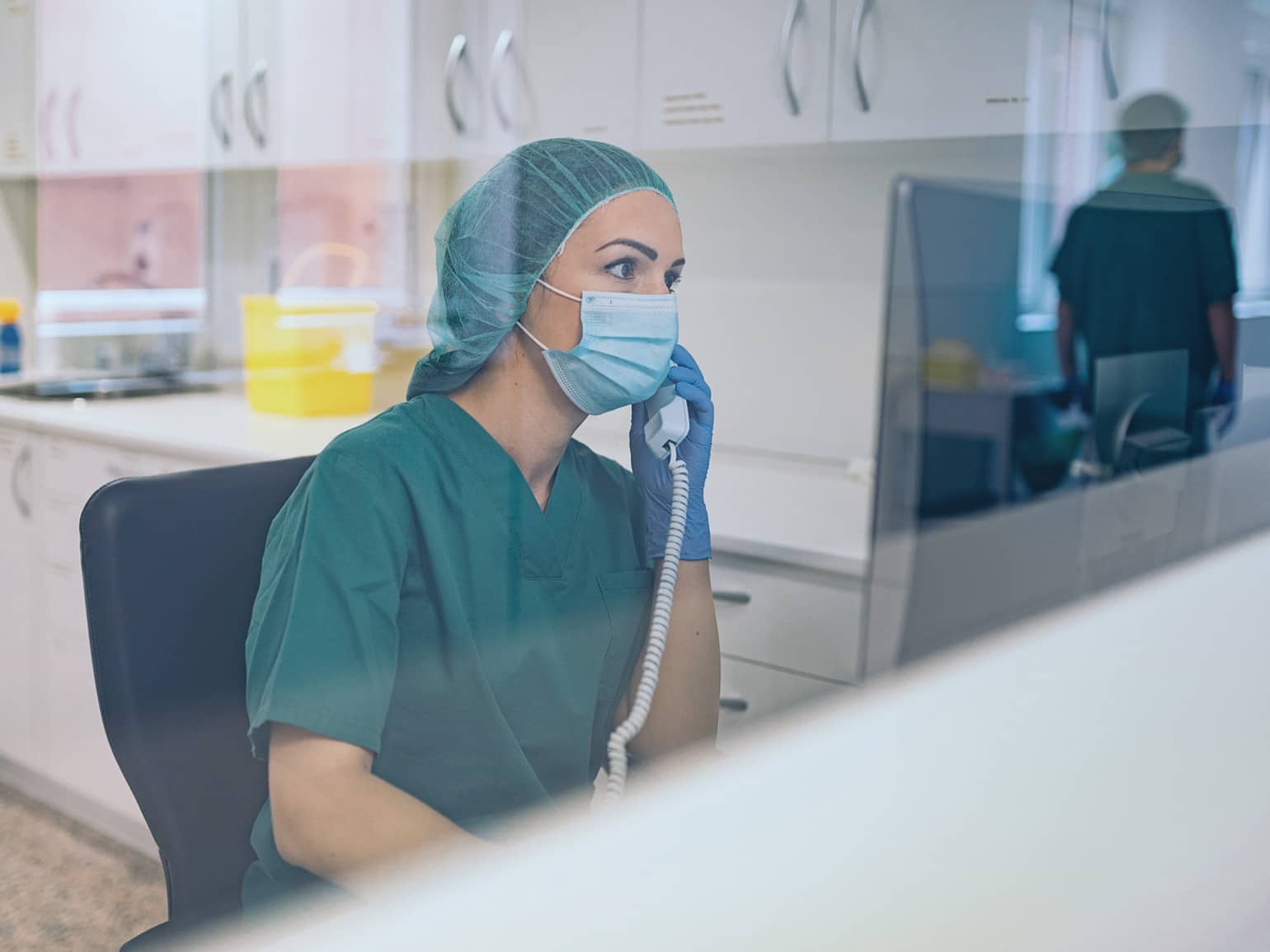 Eine Ärztin mit Maske telefoniert im Stationszimmer. Sie sitzt vor ihrem PC-Bildschirm.