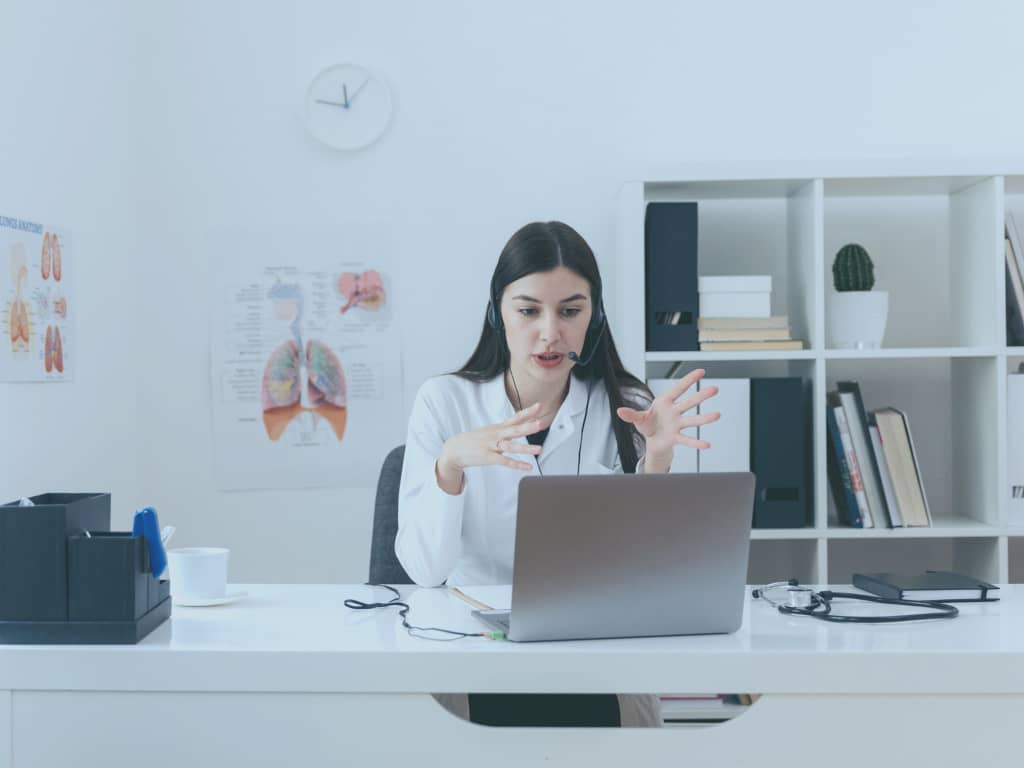 Eine Ärztin sitzt in ihrer Praxis hinter dem Schreibtisch. Sie spricht in einer Videokonferenz auf ihrem Laptop.