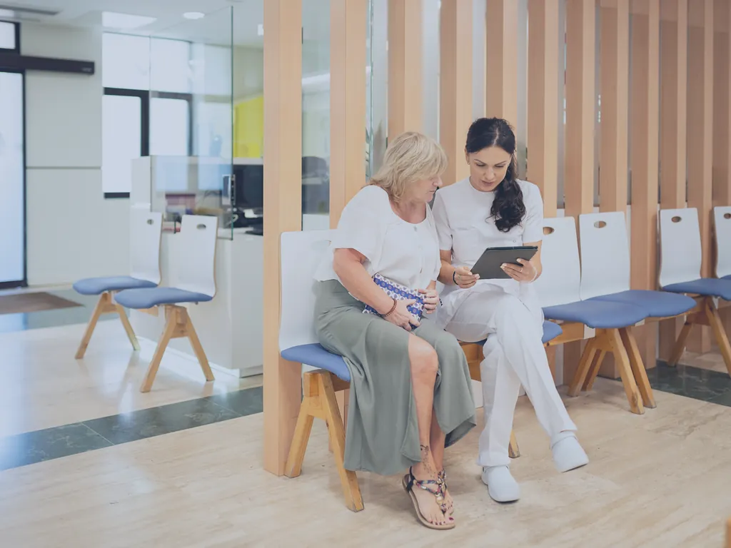 Eine Patientin und eine Klinikmitarbeiterin sehen gemeinsam auf ein Tablet im Wartebereich.